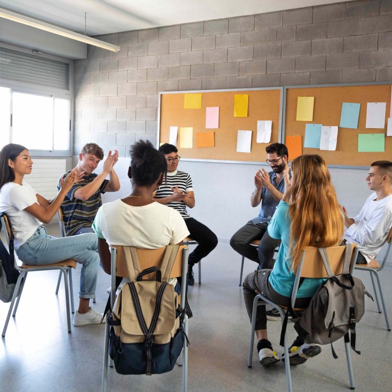 Multiracial group of high school students sitting in a circle clapping together celebrating achievement. Support group. Vertical image. Mental health concept.